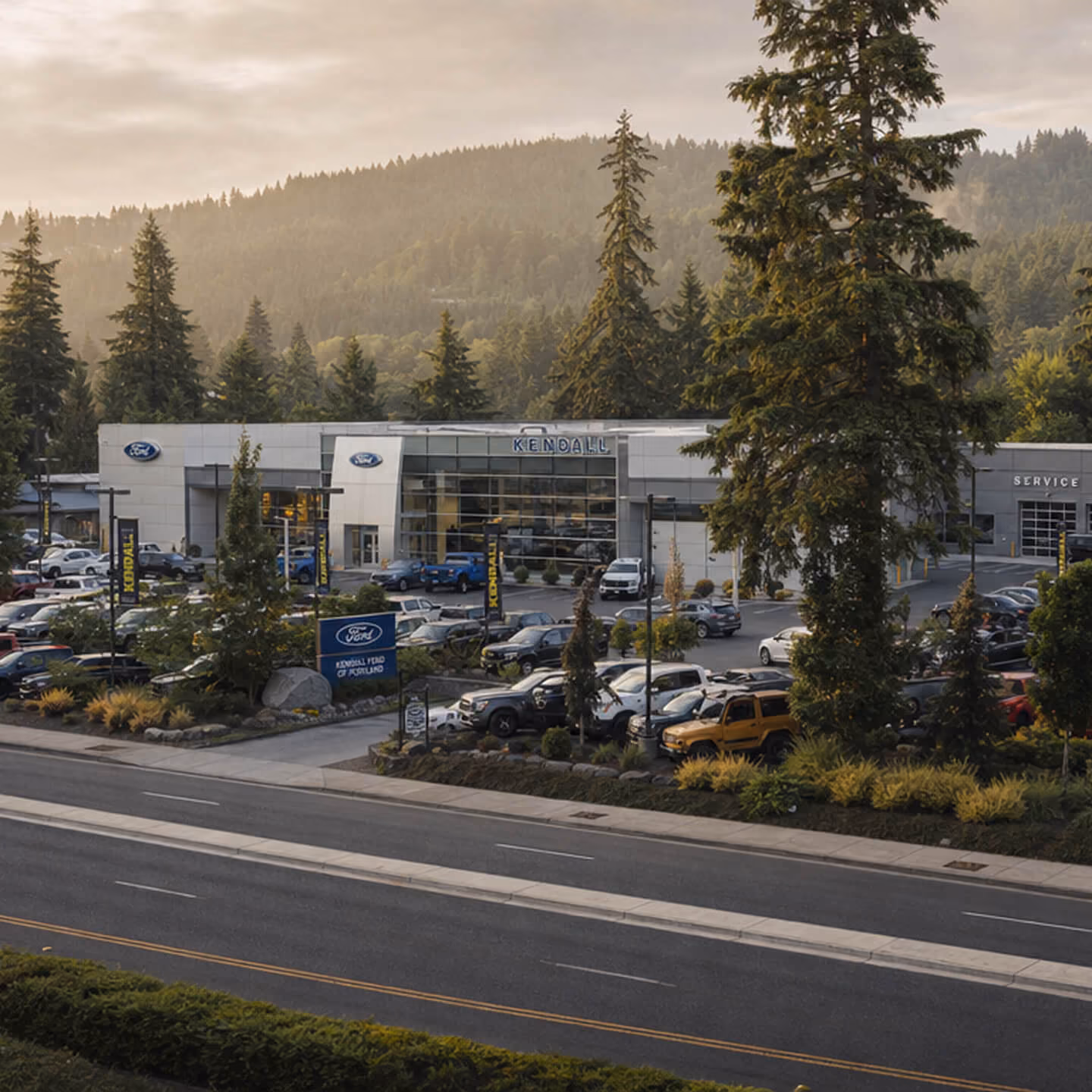 Kendall Ford car dealership with a large parking lot filled with cars, surrounded by tall trees and hills in the background during a cloudy day.