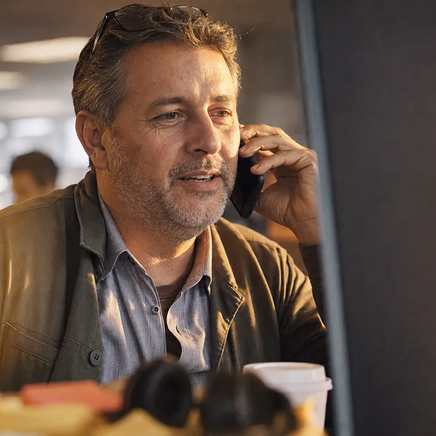 Middle-aged man with salt-and-pepper hair and stubble talking on a smartphone while looking at a computer screen, seated indoors with a coffee cup and headphones in front of him.
