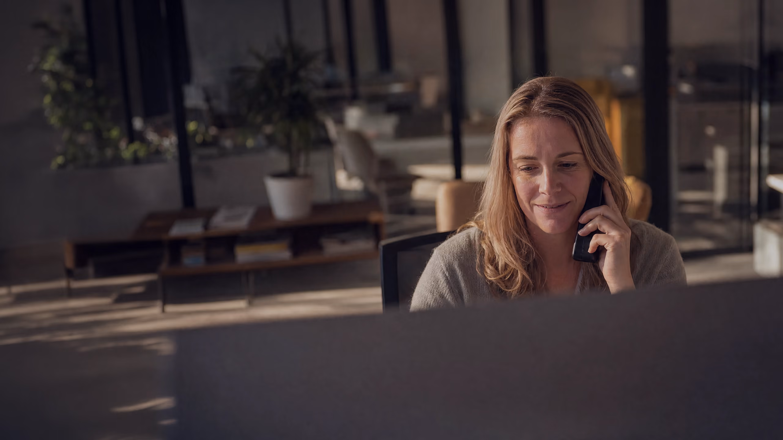 A woman with blonde hair sitting at a desk in a dimly lit office, talking on a phone with a slight smile.