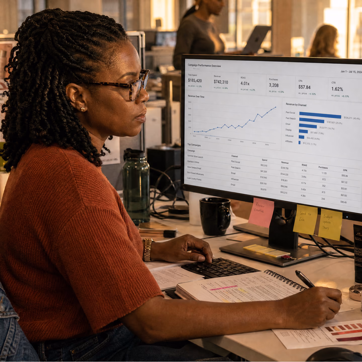 Woman with braided hair and glasses working at a desk, analyzing campaign performance data displayed on a computer screen with charts and statistics.