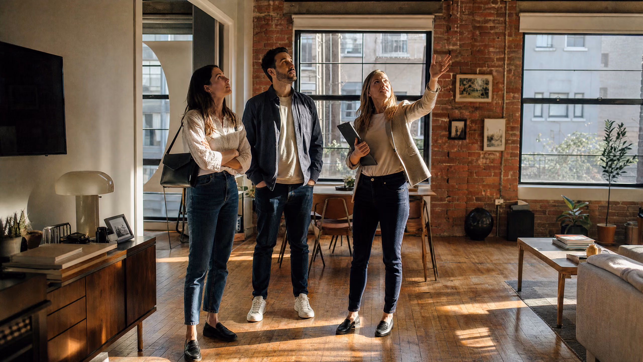 A woman showing a man and another woman features of a spacious, sunlit apartment with wooden floors and brick walls.