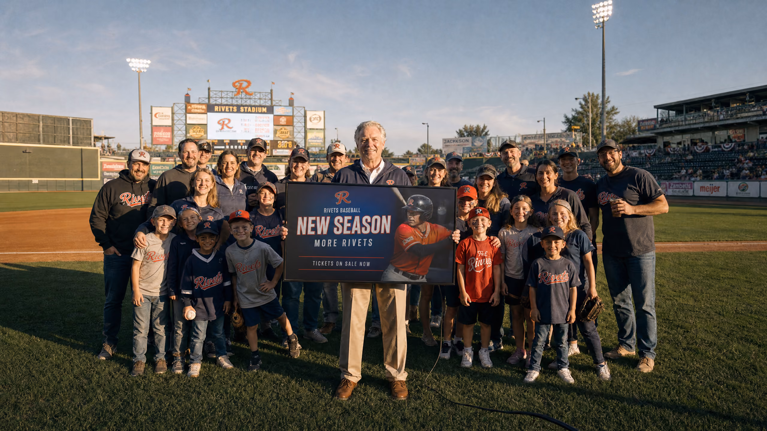A group of baseball players and supporters standing on a baseball field around an older man holding a sign that reads 'Rivets Baseball New Season More Rivets Tickets on Sale Now' at Rivets Stadium during daylight.