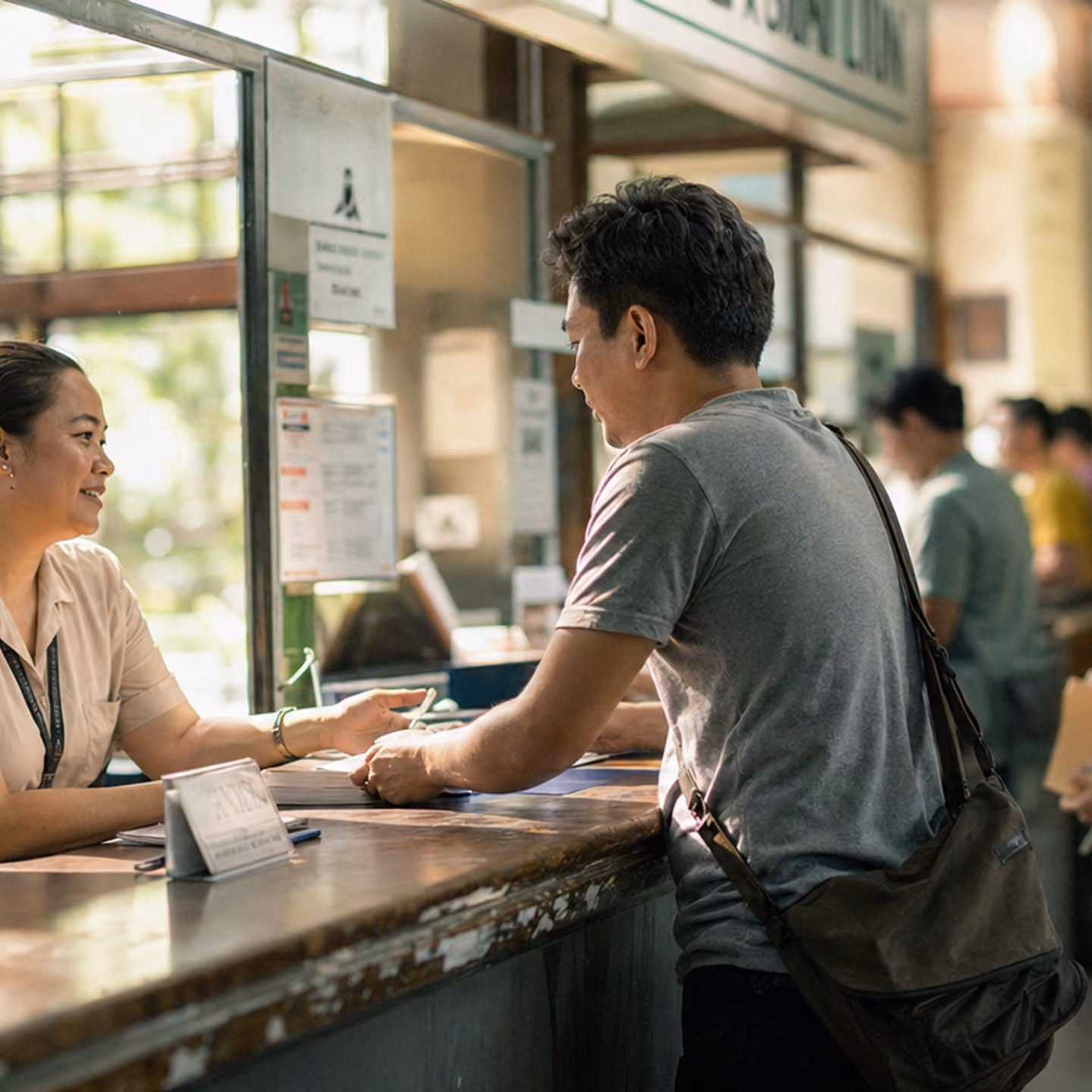 A man with a shoulder bag is talking to a smiling female employee behind a counter in an office or service center with a glass partition.