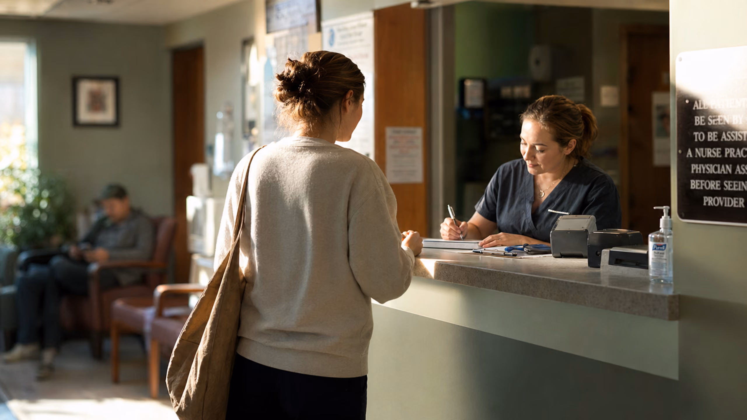A woman in a beige sweater with a shoulder bag talks to a healthcare receptionist at a clinic front desk, where the receptionist is writing on a clipboard.