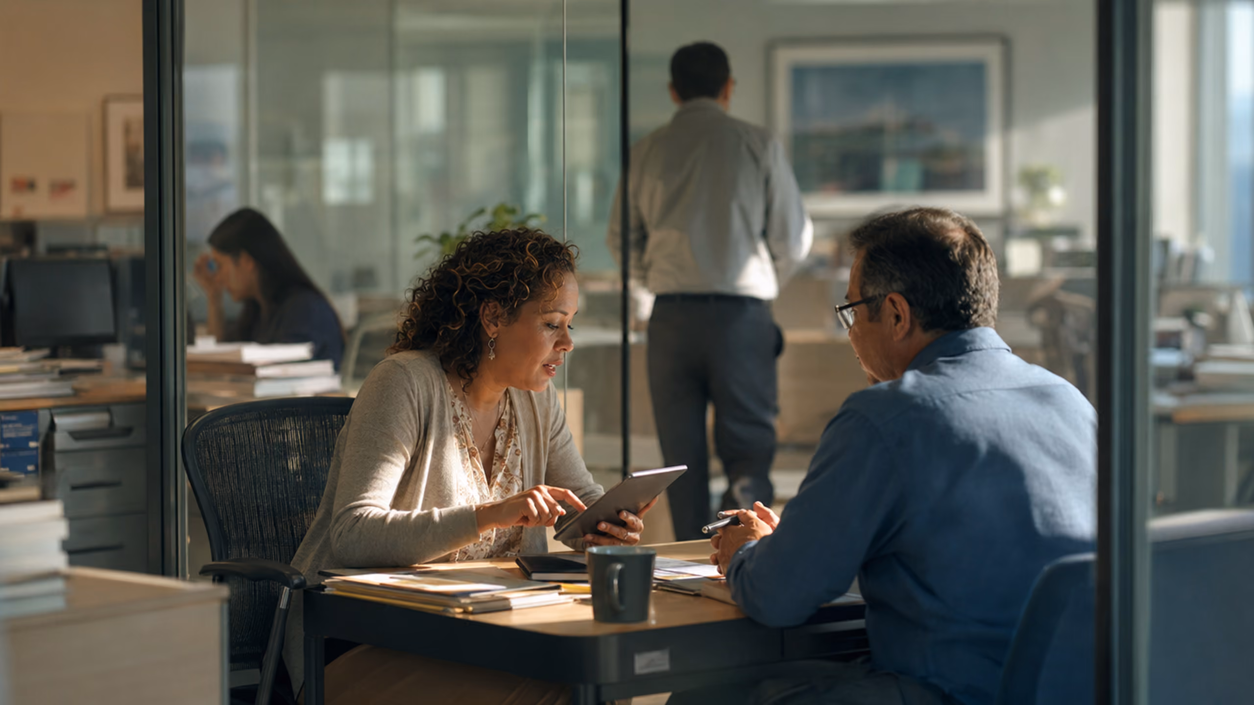 Two people engaged in a discussion across a table in an office setting, with a woman holding and pointing at a tablet, and a man listening, while another person stands in the background near a window.