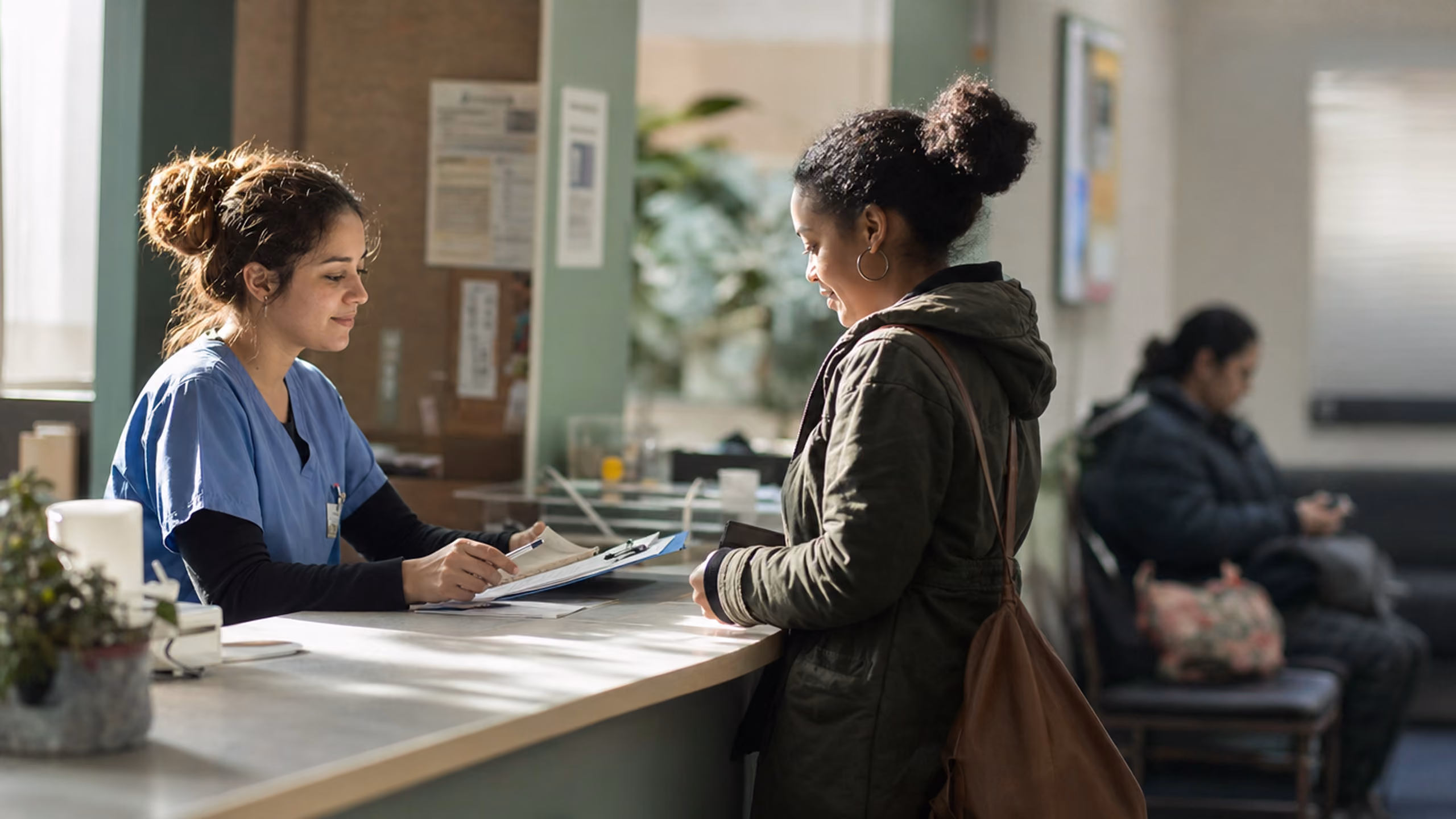 A nurse in blue scrubs smiles and consults documents on a clipboard while standing behind a reception desk talking to a woman in a dark jacket with a brown shoulder bag, in a healthcare facility waiting area with another person seated in the background.