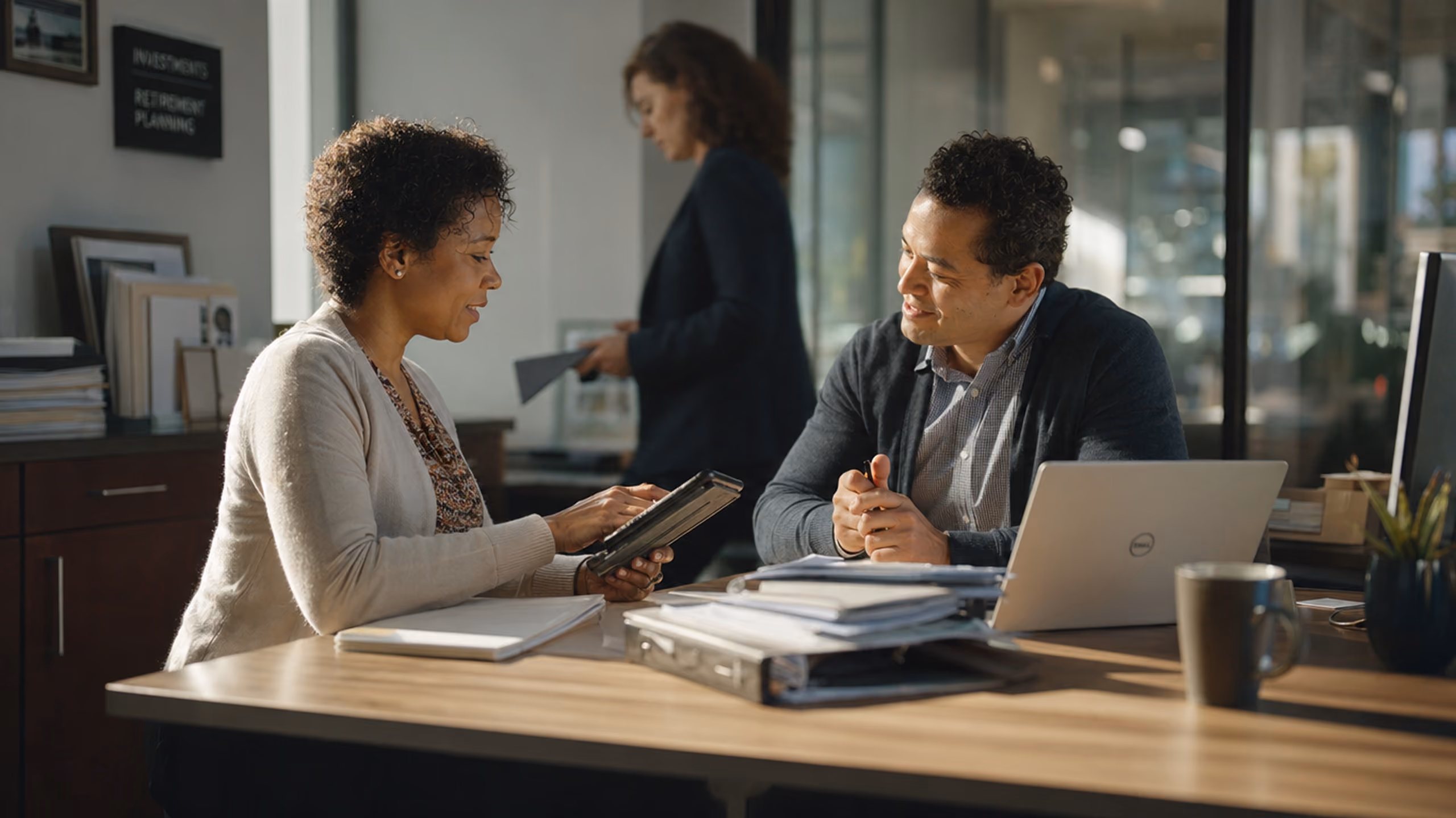 Two professionals engaged in a discussion across a desk in an office, with one person holding a tablet and the other smiling, while a third person walks in the background.