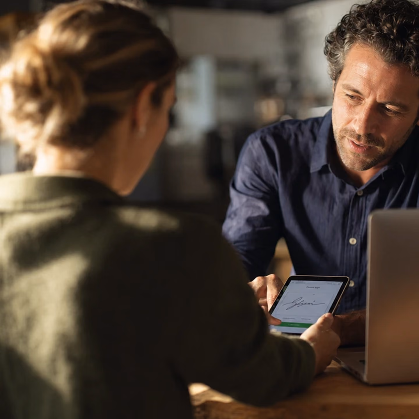 A man in a blue shirt and a woman in a green sweater sit at a table with a laptop, the man pointing at a digital signature displayed on a tablet the woman is holding.