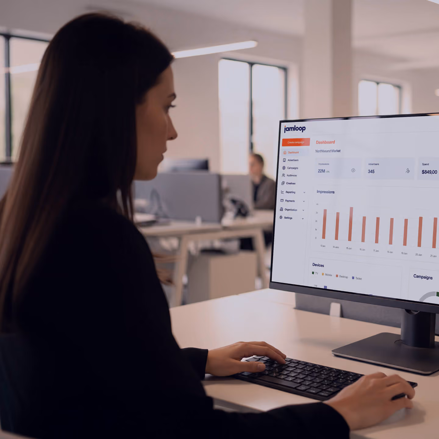 A woman with long dark hair working at a desk using a keyboard and mouse, looking at a computer monitor displaying a dashboard with charts and data in an open office setting.