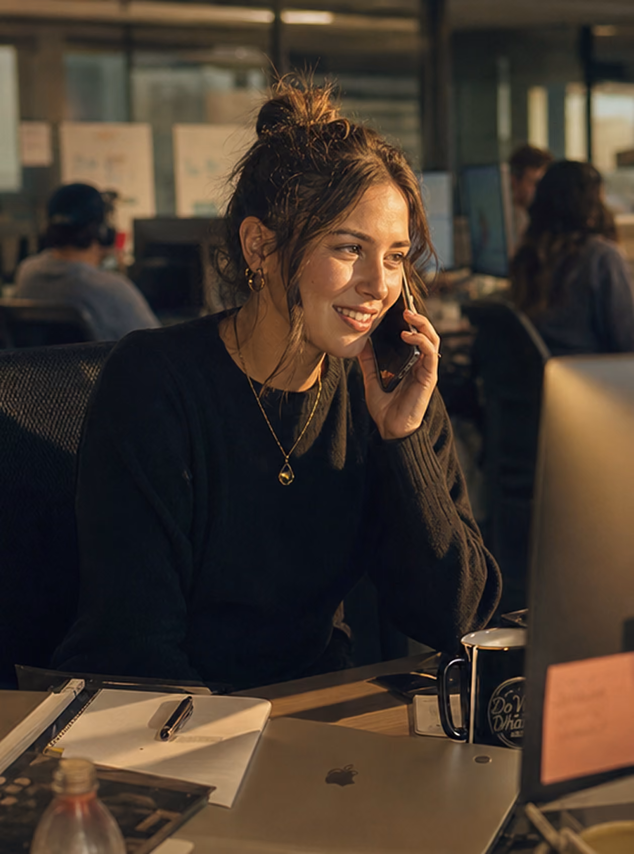 Young woman smiling and talking on a phone at a desk with a laptop, notebook, and coffee mug.