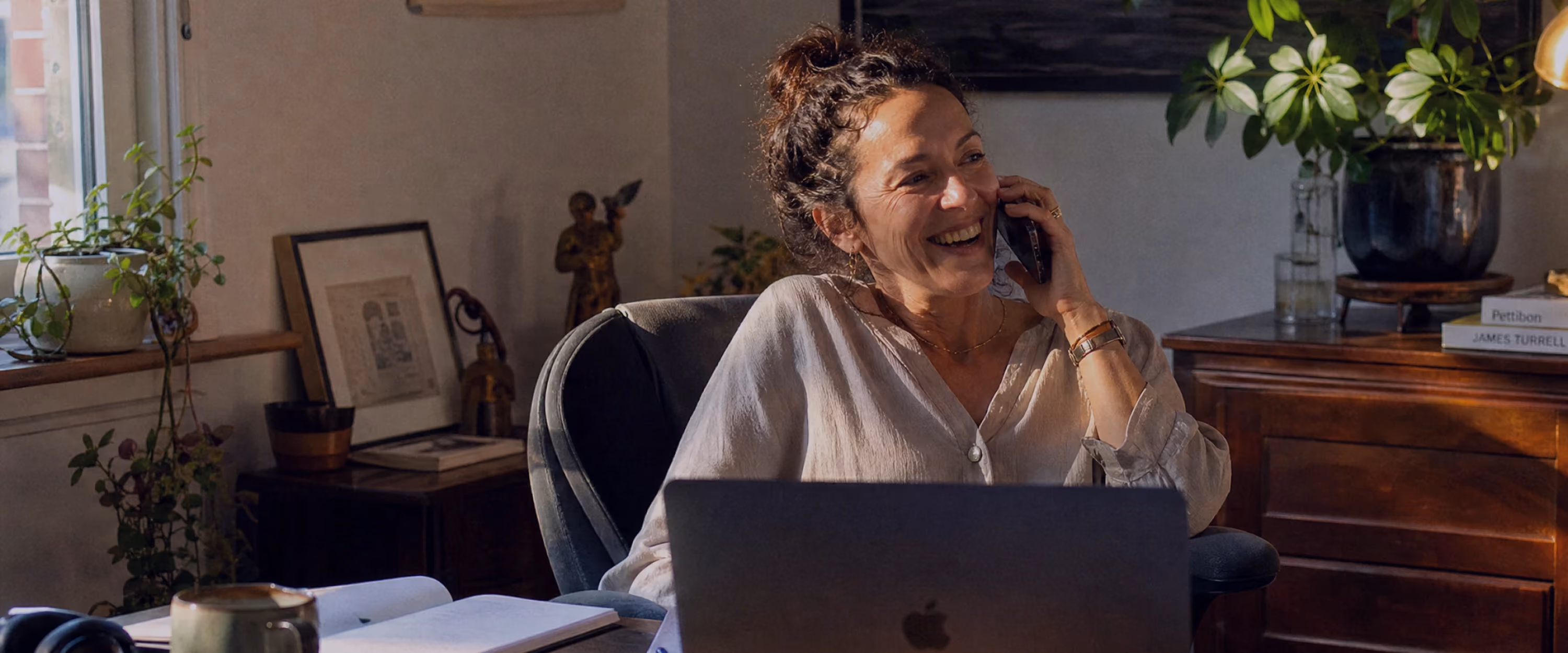 Woman laughing while talking on the phone at a cozy home desk, with a laptop, coffee mug, and notebook in front of her, surrounded by plants and vintage decor bathed in warm natural light.