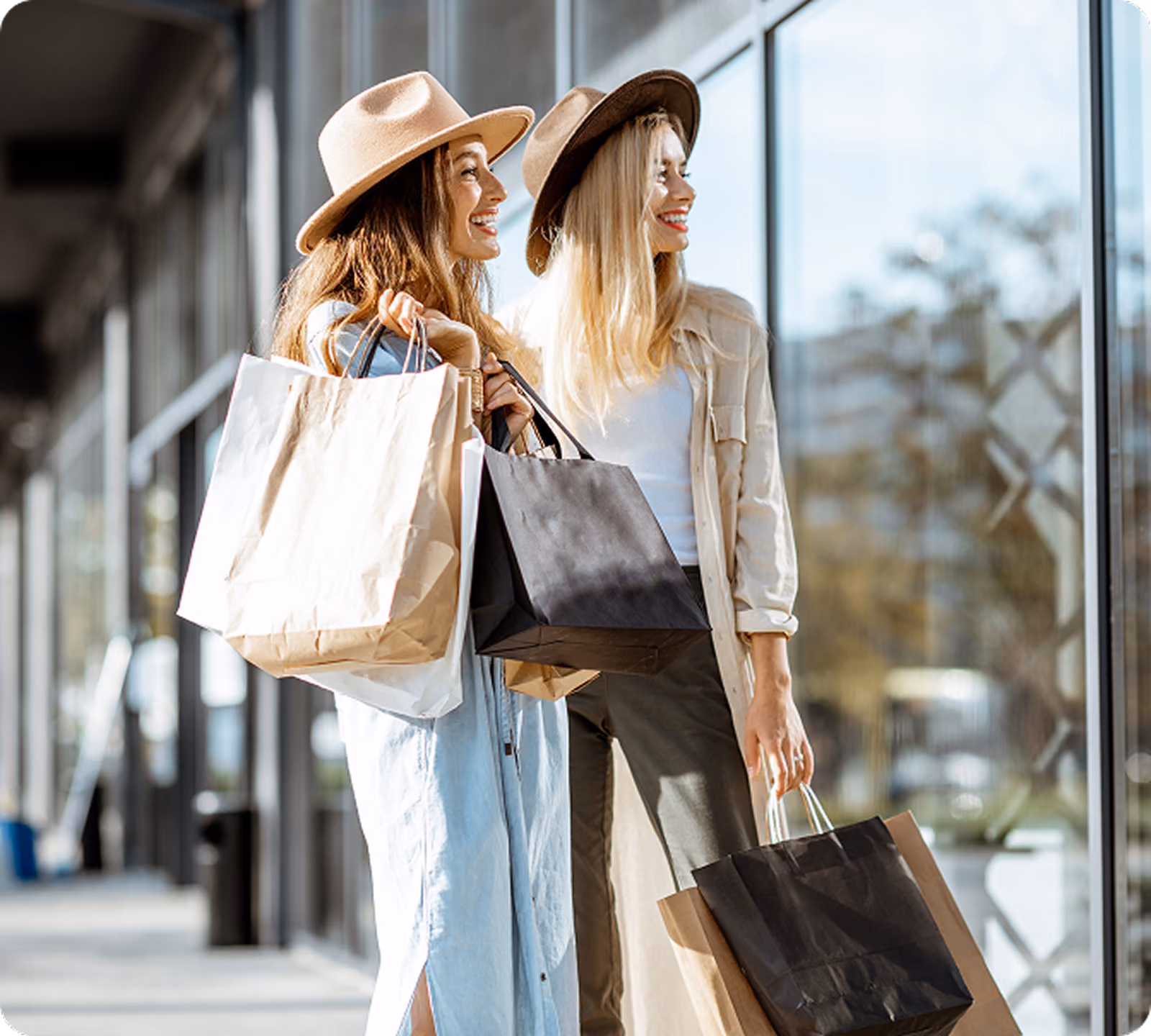 Two stylish women in hats laughing together while carrying shopping bags outside storefront windows on a sunny day.