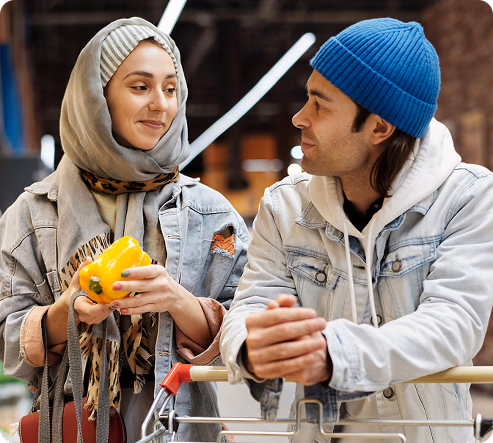 Woman in a headscarf holding a yellow bell pepper while a man leans on a shopping cart beside her, both smiling as they grocery shop together in a store.