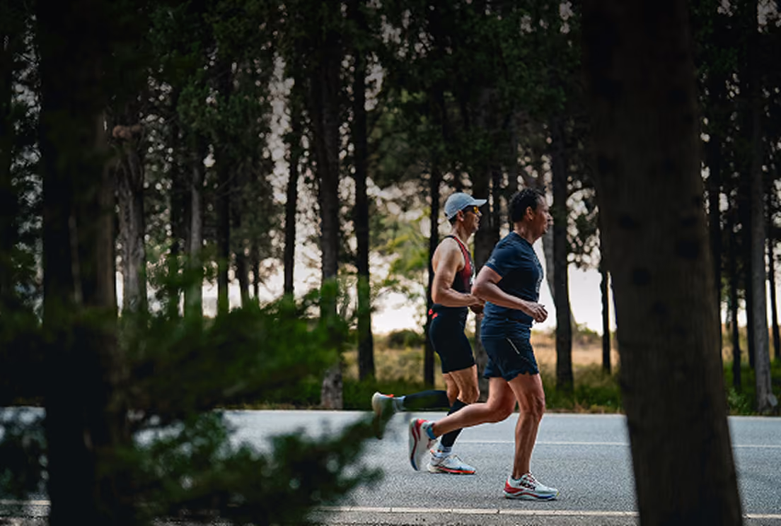 Two runners jogging along a tree-lined road, shot through silhouetted foliage in the foreground for a candid, outdoor feel.