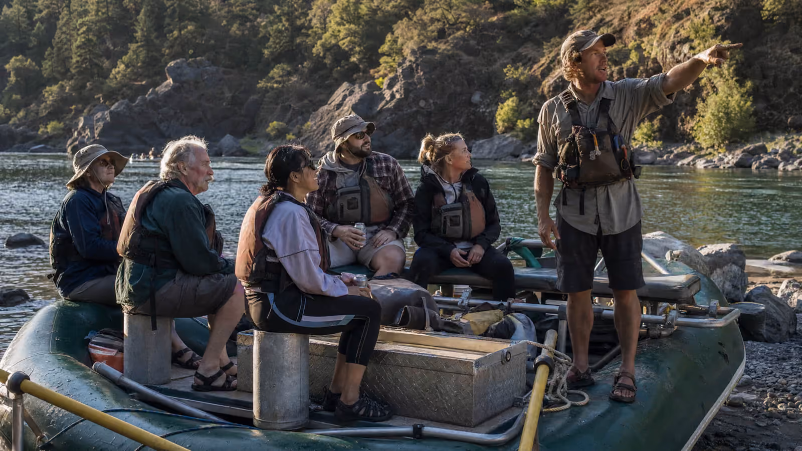 River rafting guide pointing downstream while briefing passengers seated on a raft along a forested riverbank