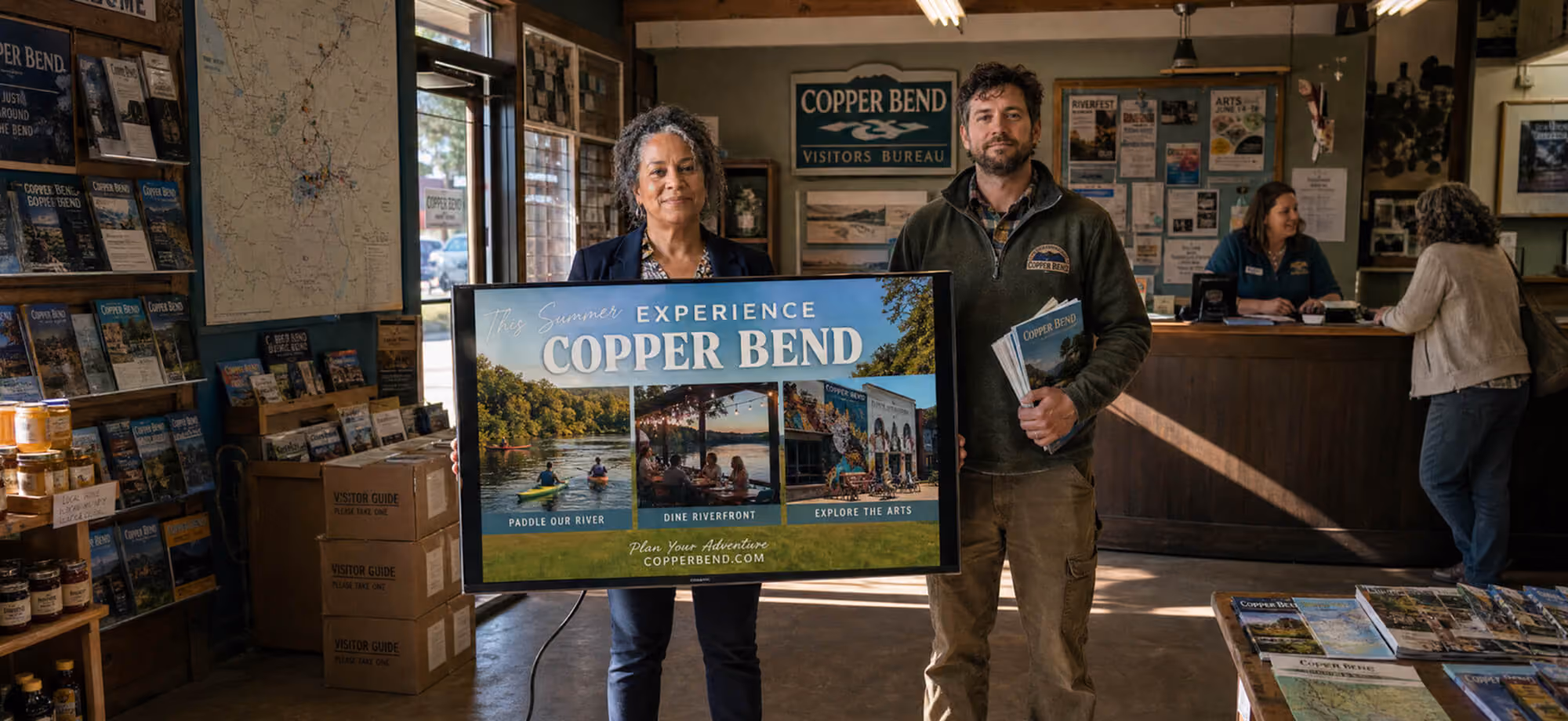 Two Copper Bend Visitors Bureau staff holding a 'This Summer Experience Copper Bend' ad display inside the visitor center