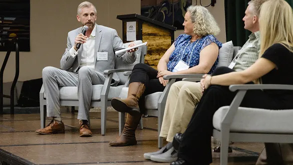 Man in gray suit speaking into a microphone during a panel discussion with three seated people.