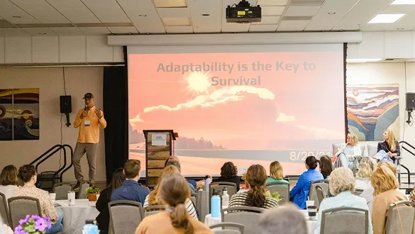 Speaker presenting on stage with slide reading 'Adaptability is the Key to Survival' to seated audience.