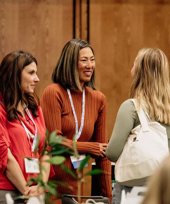 Three women at a conference smiling and greeting each other, two wearing lanyards with badges.