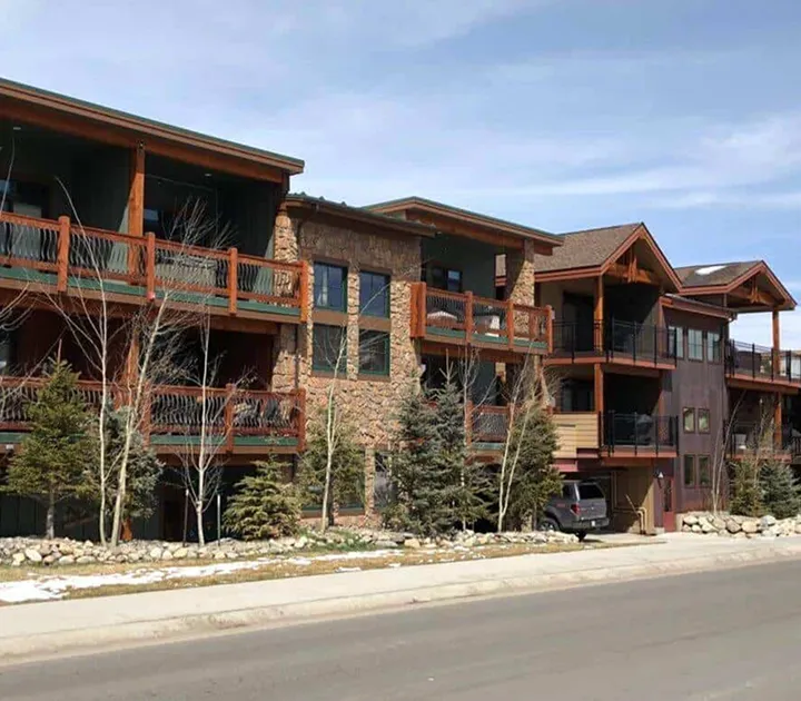 Multi-story residential building with stone and wood facade, balconies, and sparse trees in front on a clear day.