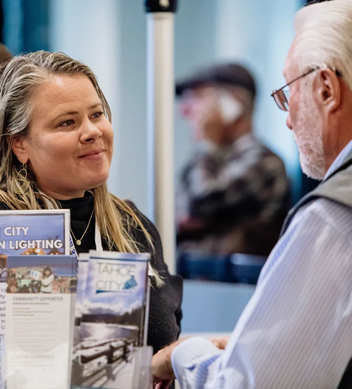 A woman hands brochures to an older man at an information desk indoors.