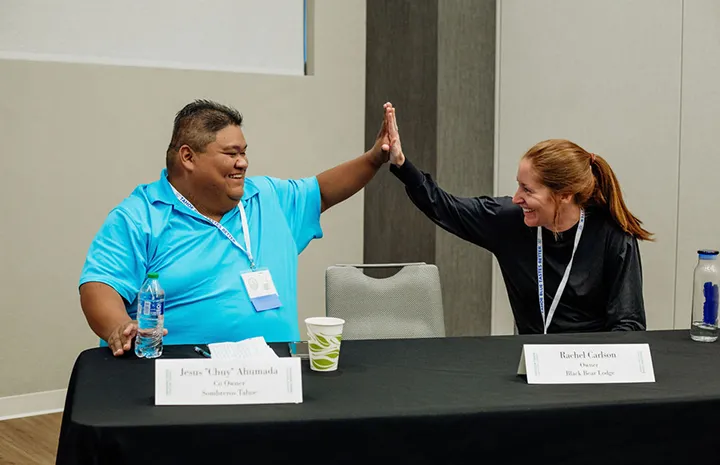 Two people sitting at a table smiling and giving each other a high five across the table.
