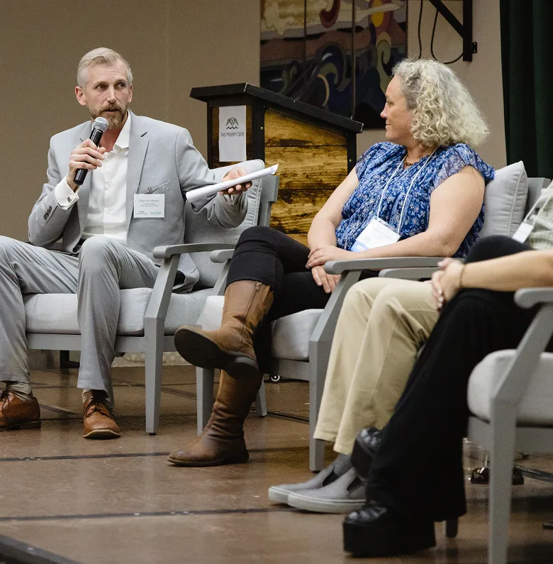 Man in gray suit speaking into microphone while seated next to three other people during a panel discussion.