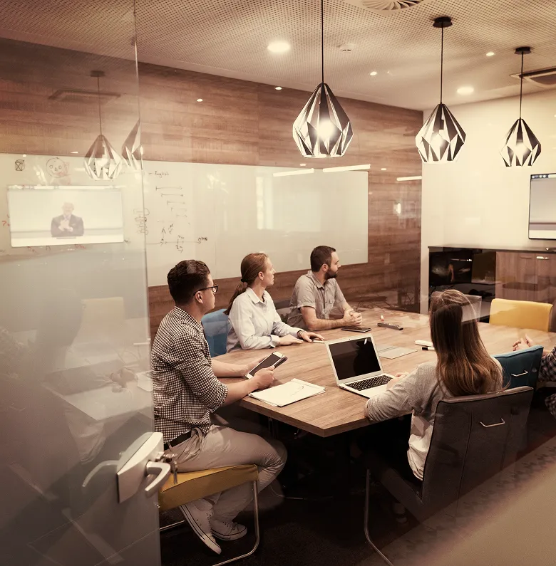 A group of four professionals sitting around a wooden conference table in a modern meeting room. They are looking forward, likely at a presentation screen or a speaker just out of frame, with a laptop and a tablet open on the table.