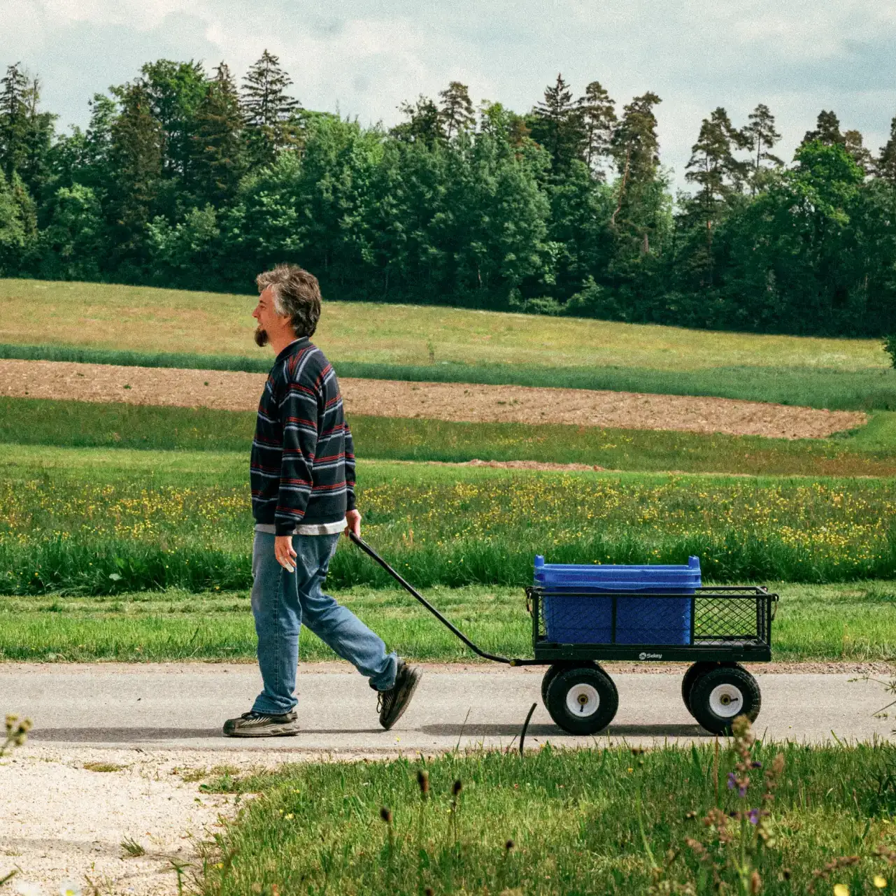 Ein Mann in Jeans und gestreiftem Pullover zieht einen Freizeitwagen mit einem blauen Behälter auf einem Weg durch eine grüne Landschaft mit Bäumen im Hintergrund.