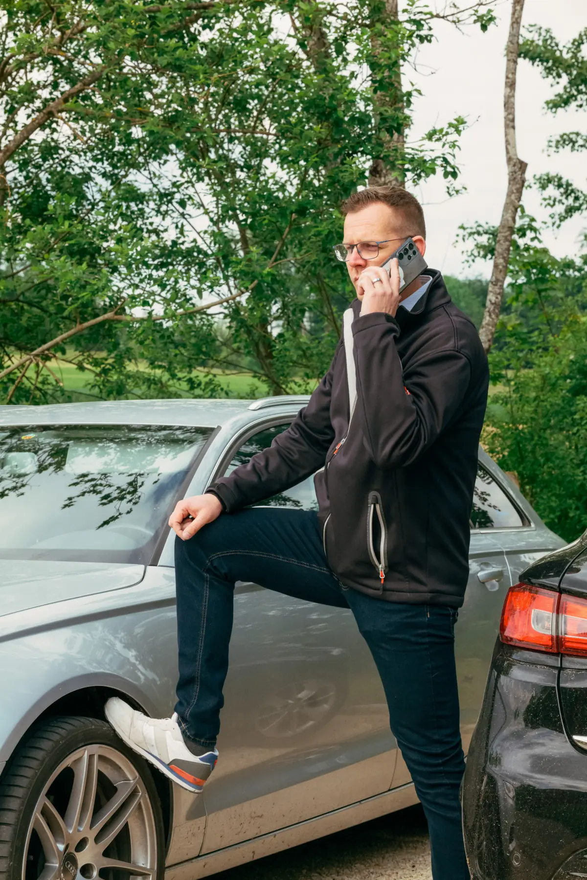 Mann in schwarzer Jacke und Jeans steht mit einem Bein auf dem silbernen Auto und telefoniert im Freien vor grünem Baum.