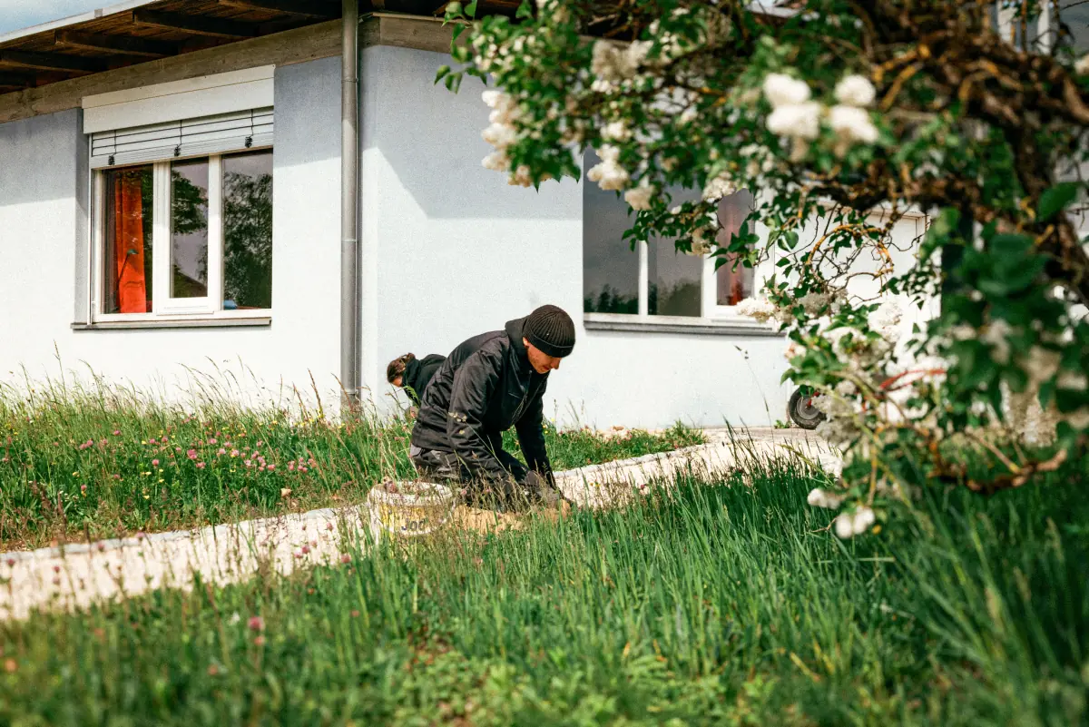Zwei Personen in dunkler Arbeitskleidung knien im Gras neben einem Fußweg vor einem hellen Gebäude und pflanzen oder jäten Unkraut in einem Garten mit blühendem Strauch im Vordergrund.
