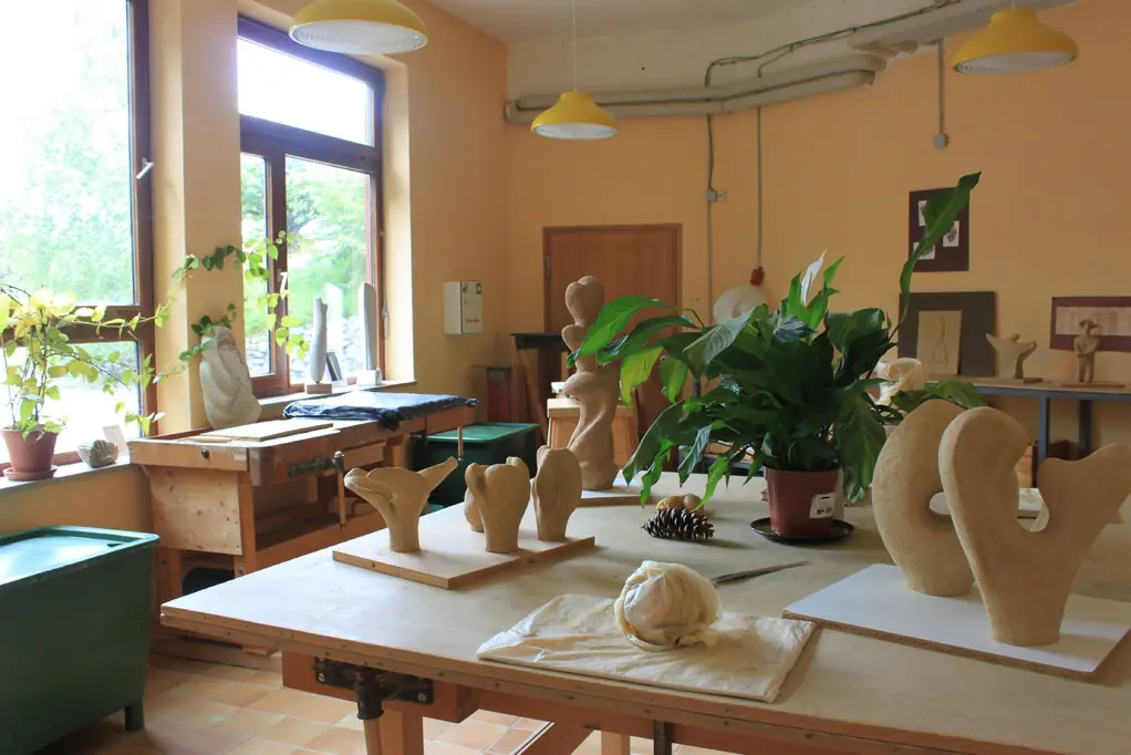Heller Kunstraum mit mehreren großen Fenstern, Holztischen mit abstrakten Skulpturen und Pflanzen, gelben Hängeleuchten und orangen Wänden.