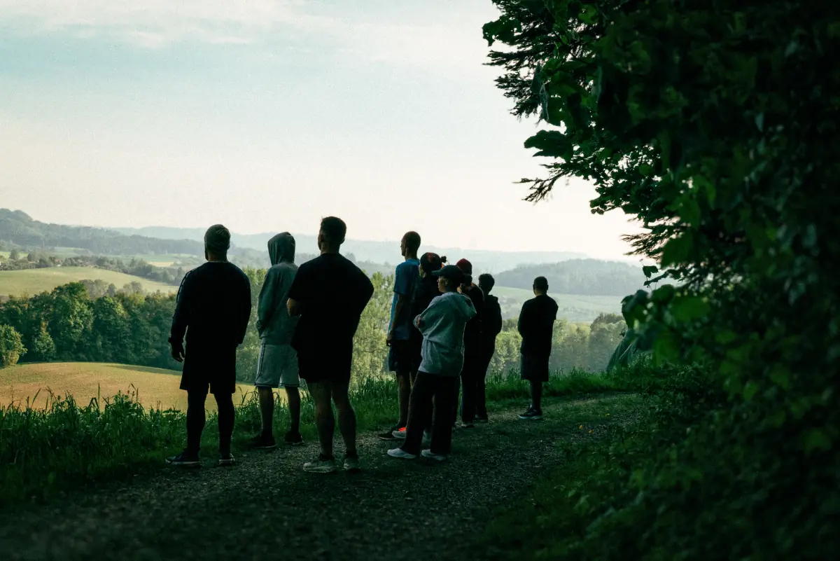 Eine Gruppe von acht Personen steht draußen auf einem Weg am Waldrand und blickt auf eine weite, hügelige Landschaft mit grünen Feldern und Bäumen.