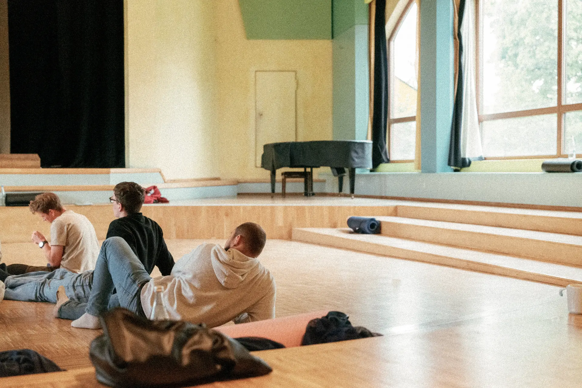 Drei junge Männer entspannt auf dem Boden eines hellen Raums mit Holzboden und großen Fenstern, im Hintergrund ein Piano und eine Treppe mit Yogamatten.