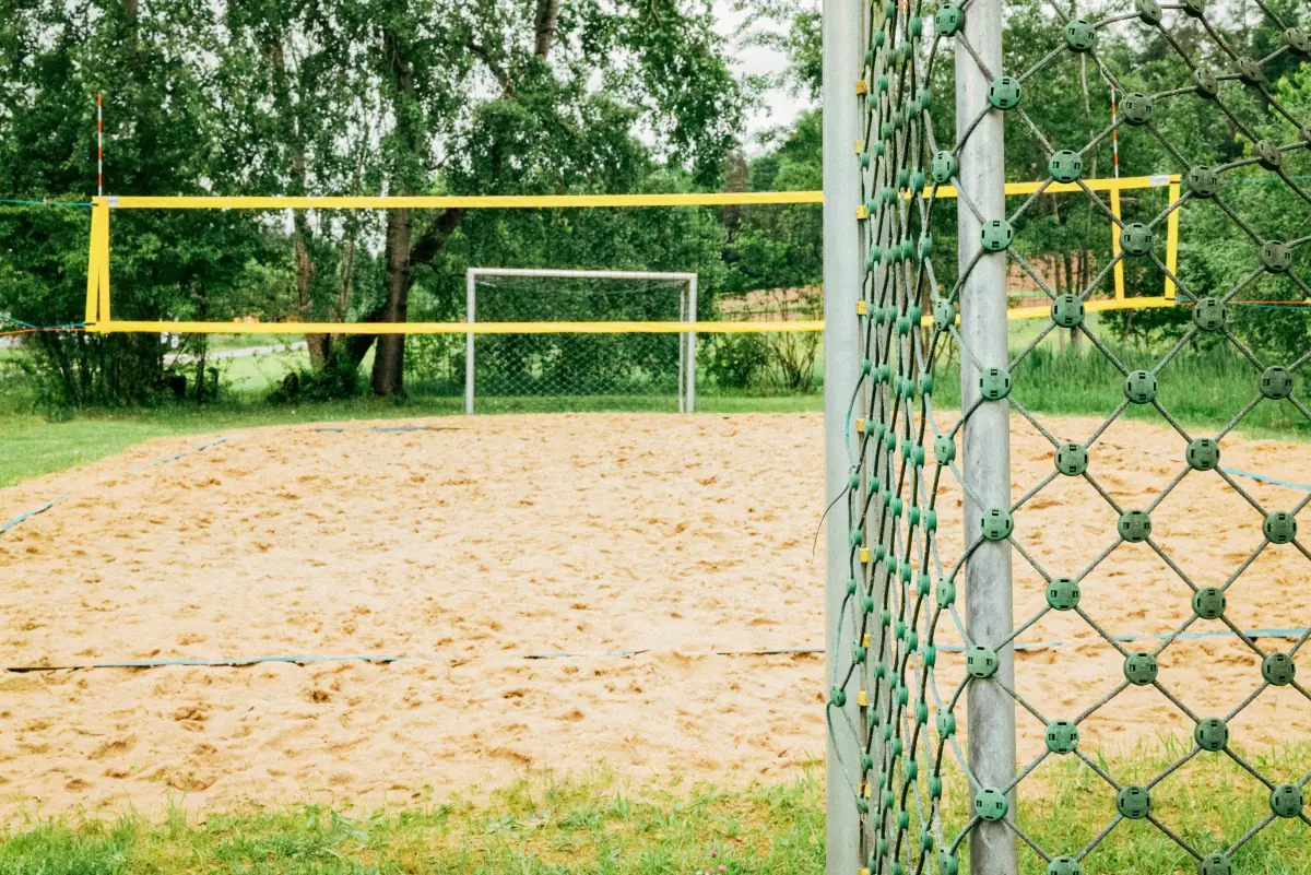 Sandvolleyballfeld mit gelbem Netz im Freien, umgeben von Grünflächen und Bäumen, im Vordergrund ist ein Drahtzaun zu sehen.