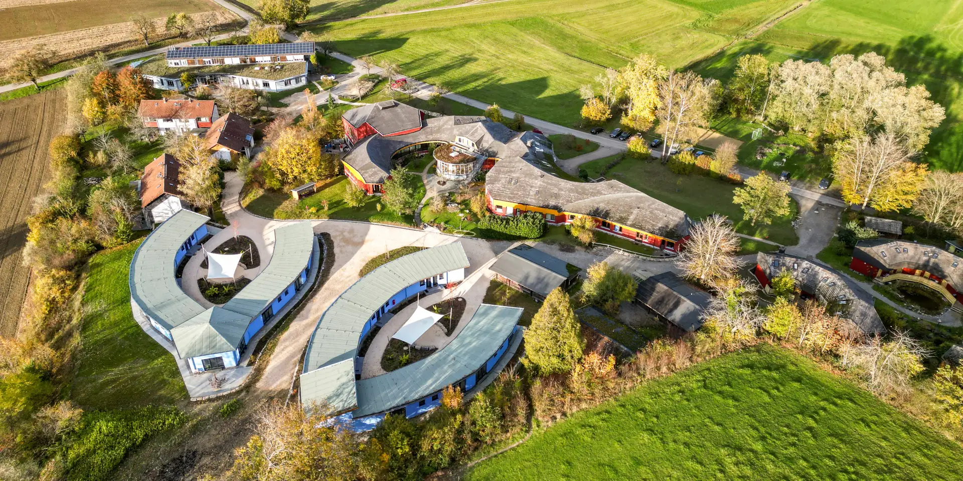 Luftaufnahme eines weitläufigen Gebäudekomplexes in ländlicher Umgebung mit grünen Wiesen, mehreren gebogenen Gebäuden mit blauen und roten Fassaden, Parkplätzen und Bäumen in herbstlichen Farben.