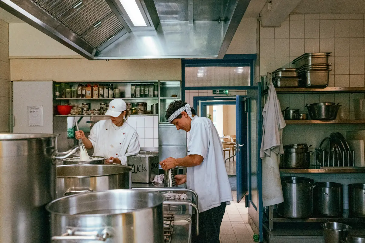 Zwei Köche in einer professionellen Küche, einer rührt in einem großen Topf, der andere bereitet Zutaten vor, umgeben von Edelstahlküchengeräten und -utensilien.