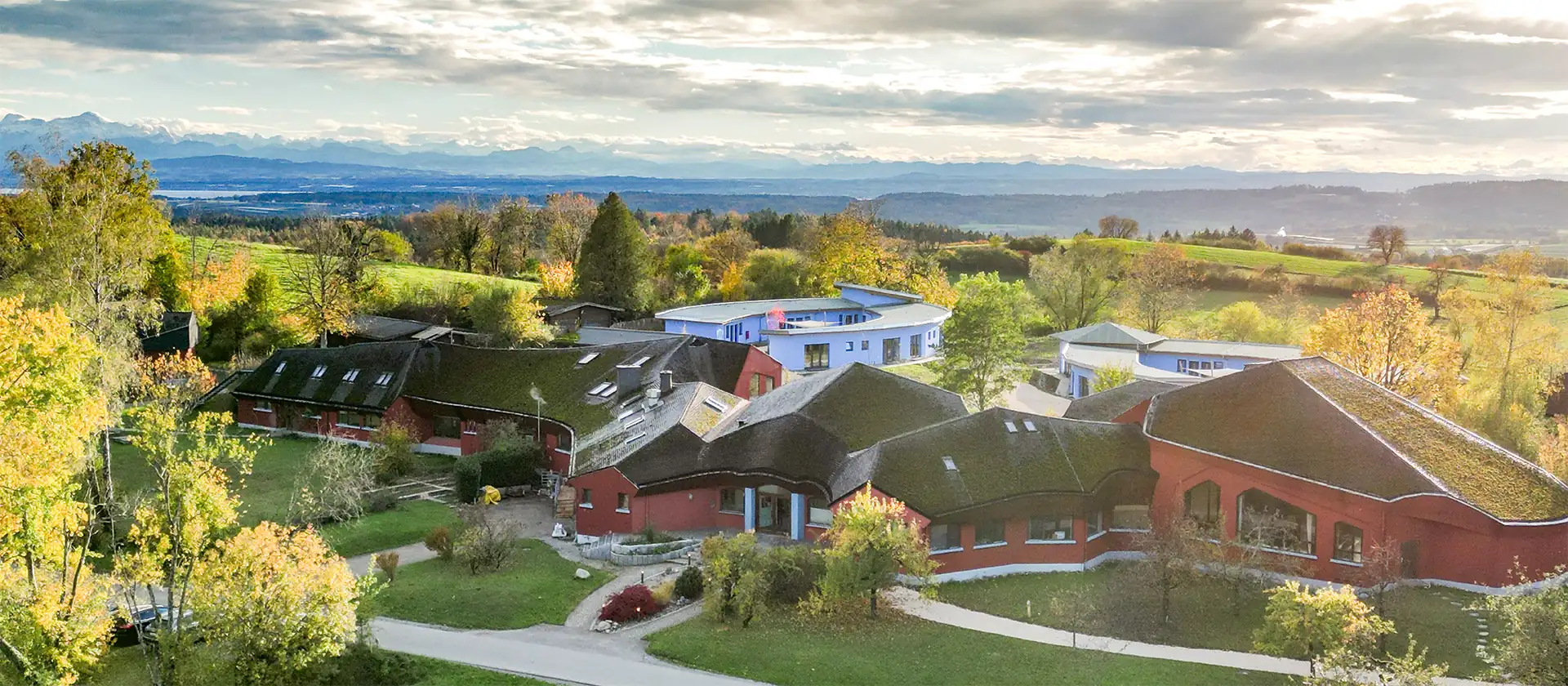 Luftaufnahme eines Gebäudekomplexes mit roten und weißen Gebäuden, umgeben von grünem Gras und herbstlichen Bäumen, im Hintergrund sanfte Hügel und eine Bergkette unter einem bewölkten Himmel.