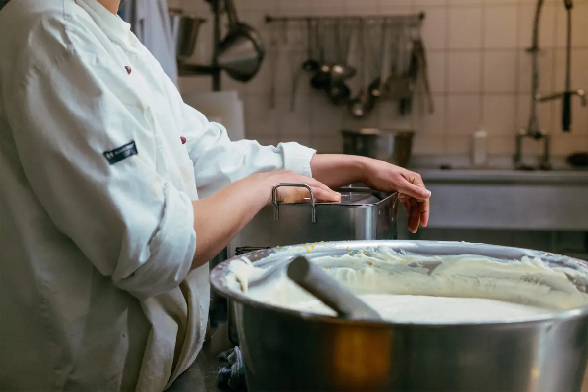 Person in weißer Kochjacke arbeitet mit einem Metallbehälter in einer professionellen Küche, im Vordergrund großer Behälter mit cremigem weißen Inhalt und Rührgerät.