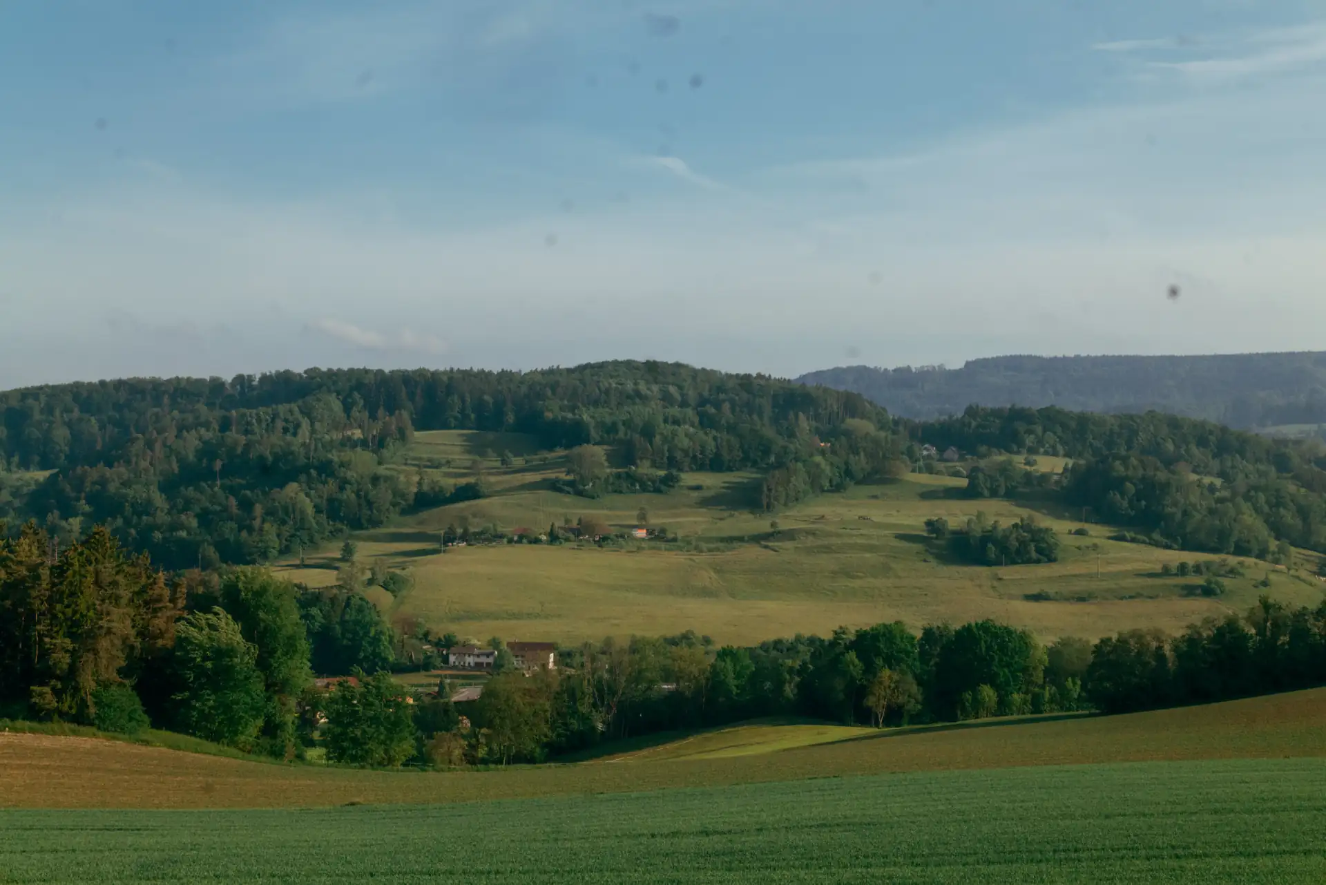 Landschaft mit grünen Feldern im Vordergrund und bewaldeten Hügeln im Hintergrund unter einem blauen Himmel mit wenigen Wolken.