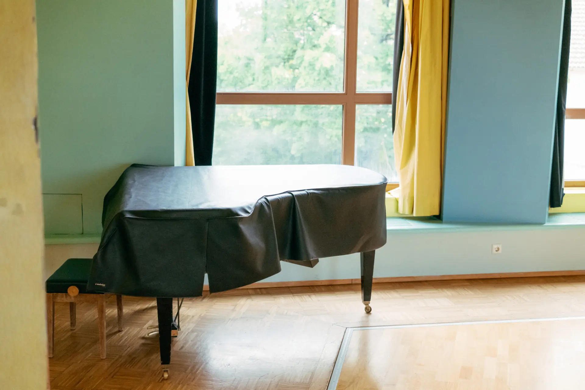 Ein Flügelklavier mit schwarzer Abdeckung steht in einem hellen Raum vor einem großen Fenster mit gelben Vorhängen.
