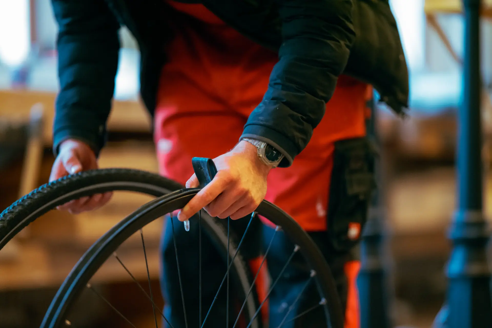 Person in roter Arbeitskleidung und schwarzer Jacke wechselt oder repariert einen Fahrradreifen und hält dabei einen Fahrradreifen und eine Felge in den Händen.