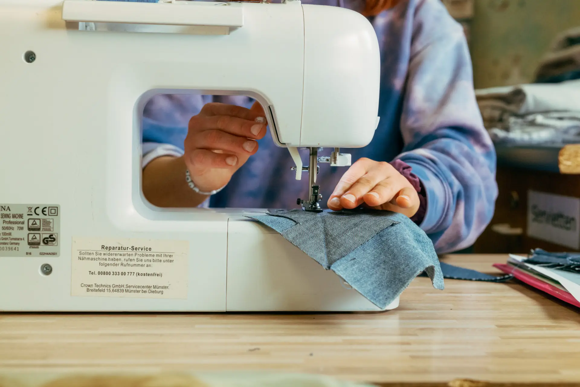 Person näht grau melierten Stoff an einer weißen Nähmaschine auf einem Holztisch, mit Händen, die den Stoff führen und das Nähwerkzeug einstellen.