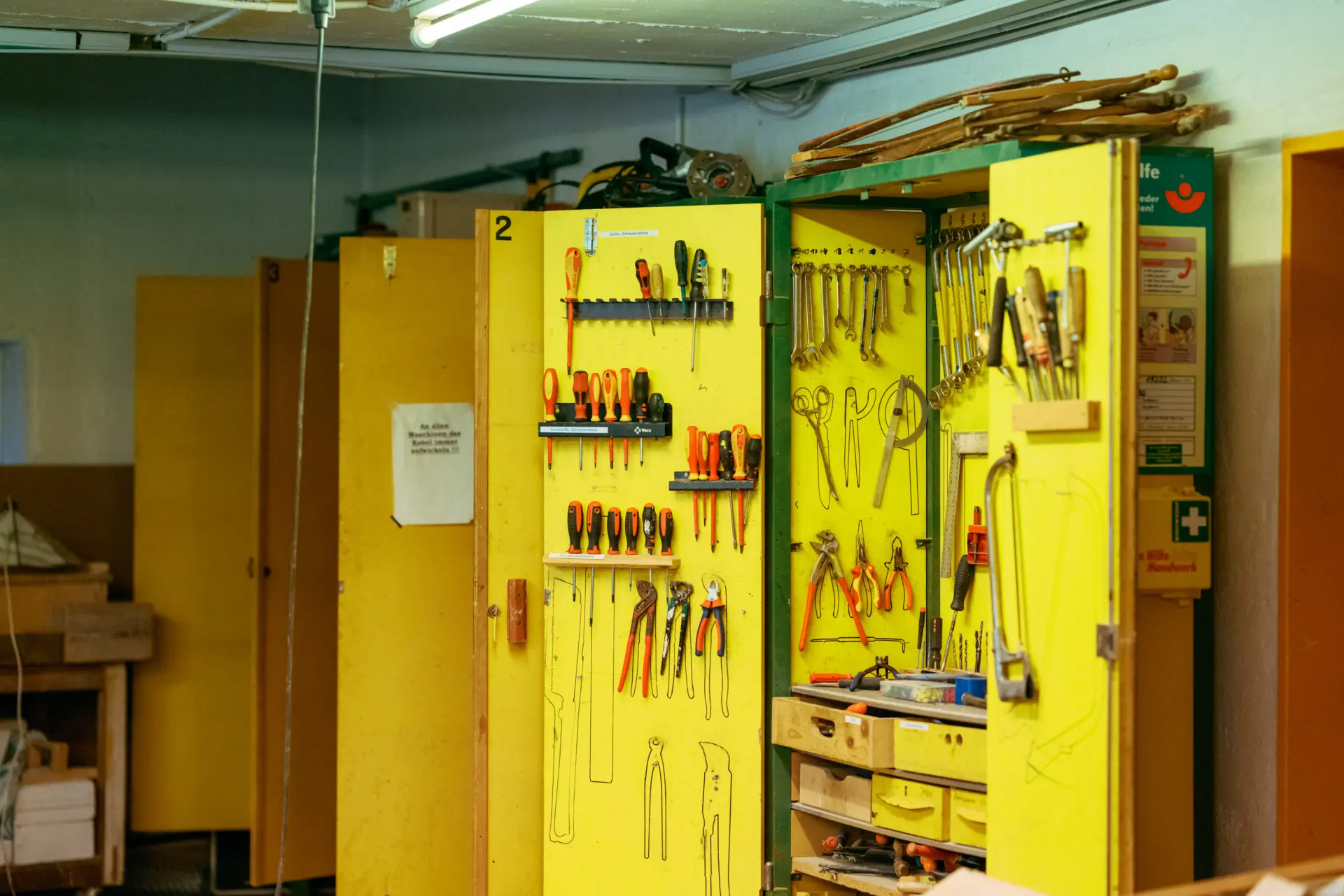 Gelber Werkstattschrank mit offenstehenden Türen, an denen verschiedene Schraubendreher, Zangen, Schraubenschlüssel und andere Werkzeuge übersichtlich hängen, daneben mehrere geschlossene gelbe Schränke in einem Keller- oder Werkraum.