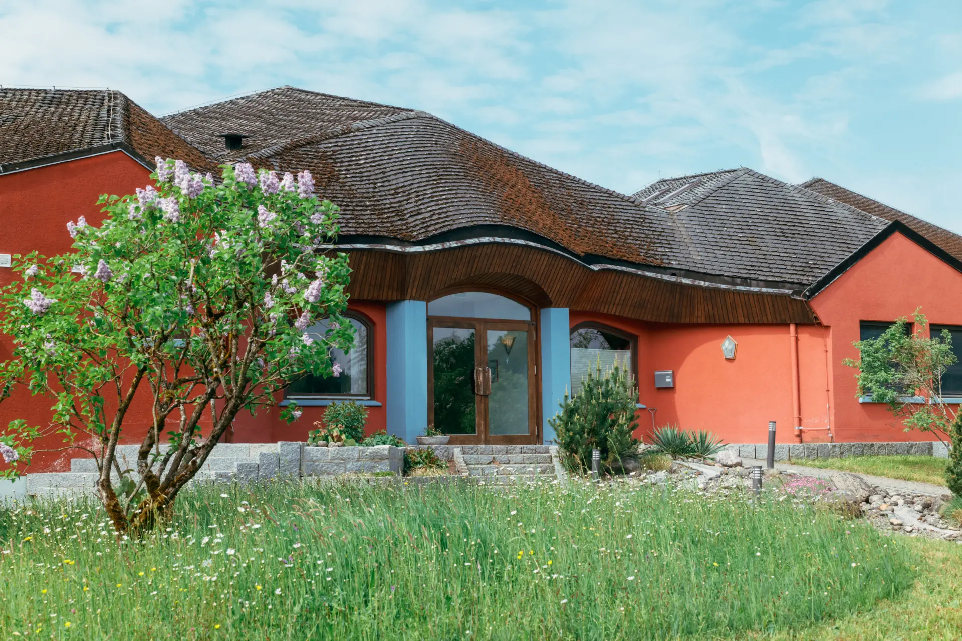 Rotes Haus mit gewelltem Schindeldach, blühendem Fliederbusch links, grüner Wiese mit Wildblumen im Vordergrund und blau umrahmtem Eingang mit Holz-Glas-Tür.