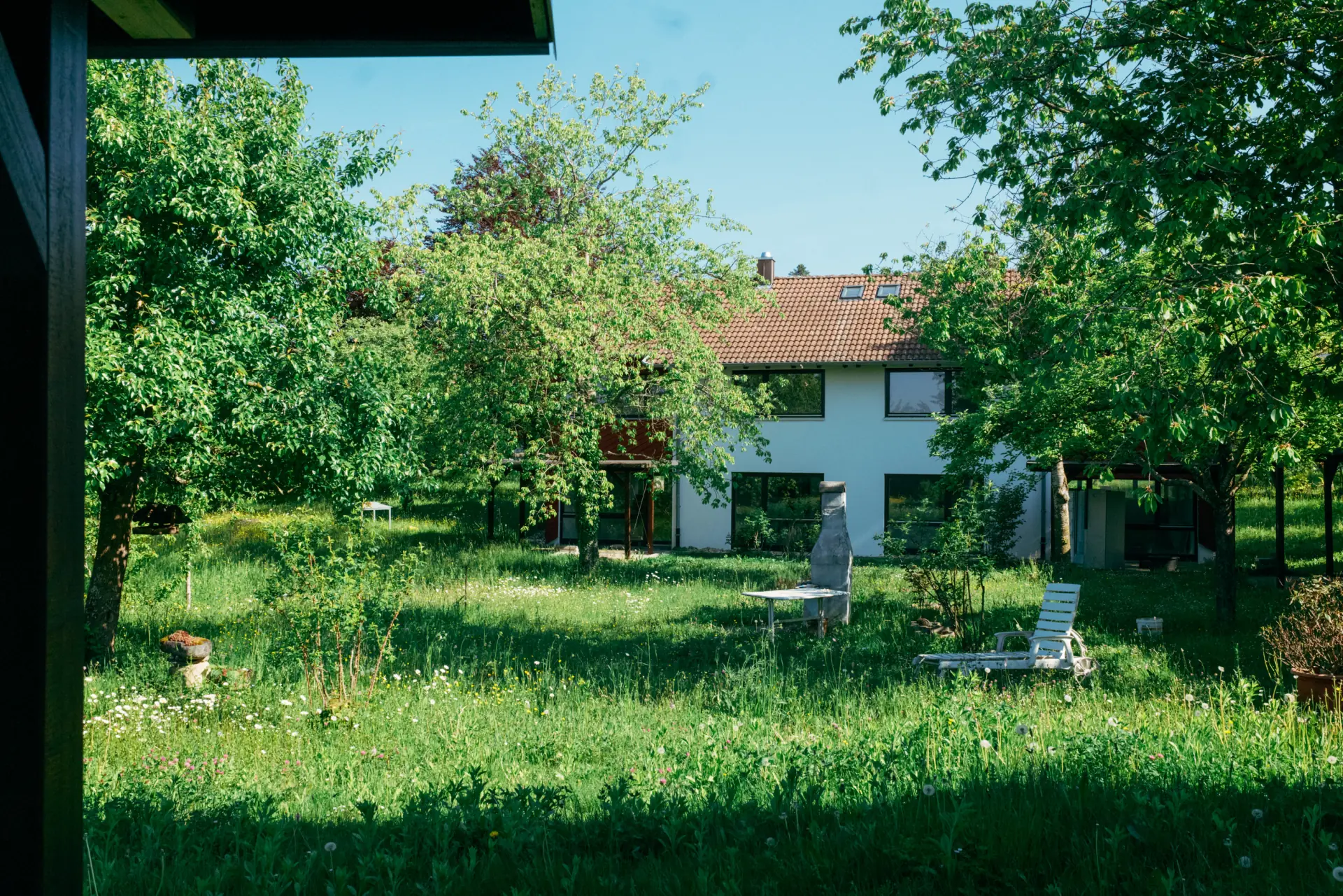 Haus mit weißer Fassade und rotem Ziegeldach hinter einem großen, grünen Garten mit hohem Gras, mehreren Bäumen, einem weißen Gartenstuhl und einem kleinen Tisch.