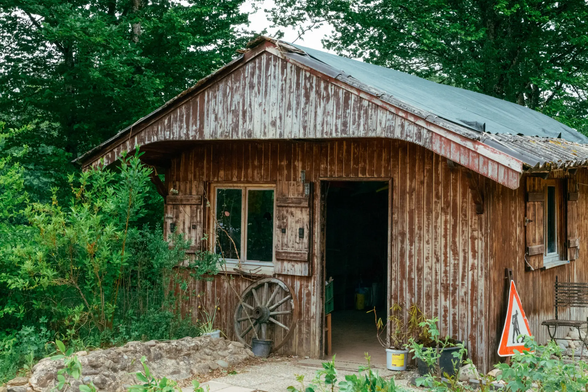Verwitterte Holzhütte mit offenem Eingang und Fenstern mit Klappläden, umgeben von grünen Büschen und Bäumen, mit einem alten Wagenrad an der Vorderseite und einem Baustellenschild rechts.