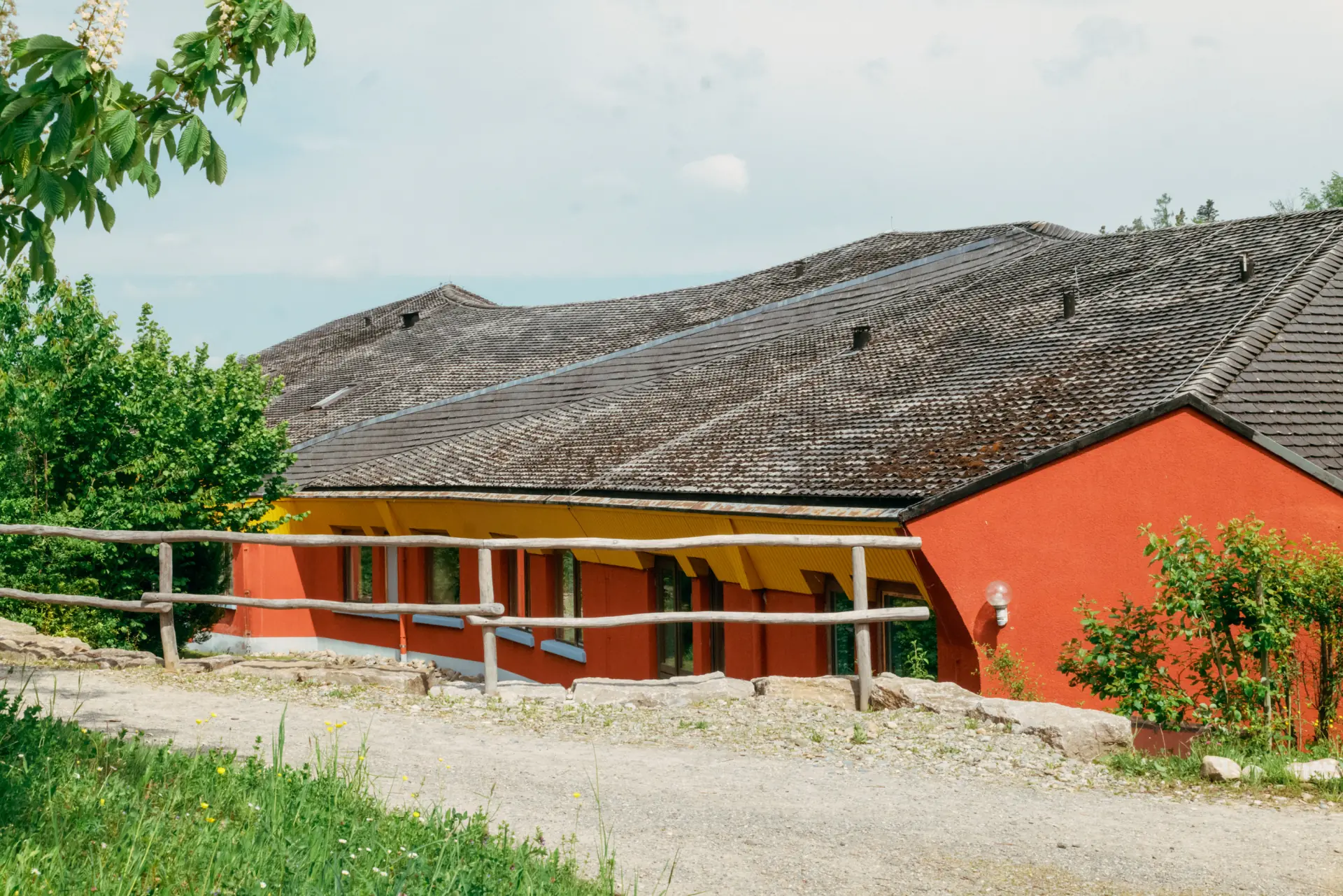 Ein langgestrecktes Gebäude mit einem großen, schrägen Dach aus dunklen Dachziegeln, orangefarbenen Wänden und gelbem Vordach, umgeben von grünen Büschen und Bäumen; im Vordergrund ein Kiesweg mit hölzernem Geländer.