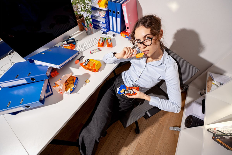 Person at a cluttered office desk eating snacks between blue binders