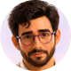 Hombre joven con cabello oscuro, barba y gafas marrones, fondo circular rosa.